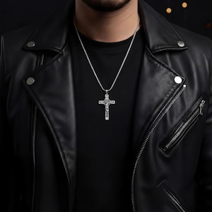May include: A silver cross pendant necklace. The cross features a detailed depiction of Jesus Christ. The necklace is worn over a black shirt and a black leather jacket. The chain is a thin, silver snake chain.