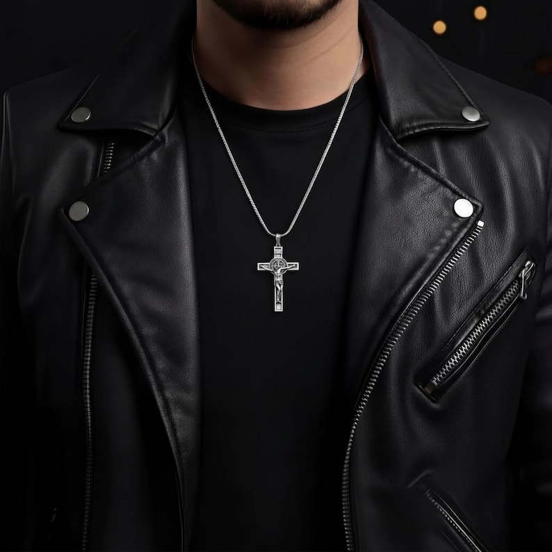 May include: A silver cross pendant necklace. The cross features a detailed depiction of Jesus Christ. The necklace is worn over a black shirt and a black leather jacket. The chain is a thin, silver snake chain.