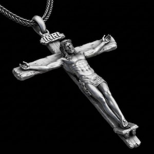 May include: A silver-colored pendant necklace featuring a detailed depiction of Jesus on a cross. The cross has a rustic, wooden appearance, and the figure is intricately sculpted. The necklace chain is a woven design, and the background is black.