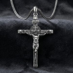 Puede incluir: Collar con colgante de cruz plateada. El colgante presenta una detallada representaci&oacute;n de Jes&uacute;s en la cruz, con intrincados detalles en la cruz. El collar se muestra sobre un fondo oscuro y texturizado.