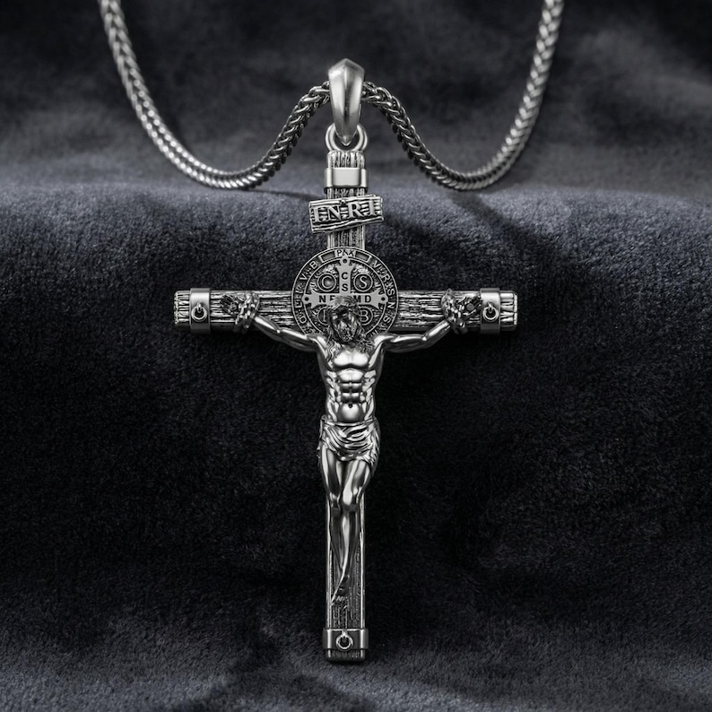Puede incluir: Collar con colgante de cruz plateada. El colgante presenta una detallada representaci&oacute;n de Jes&uacute;s en la cruz, con intrincados detalles en la cruz. El collar se muestra sobre un fondo oscuro y texturizado.