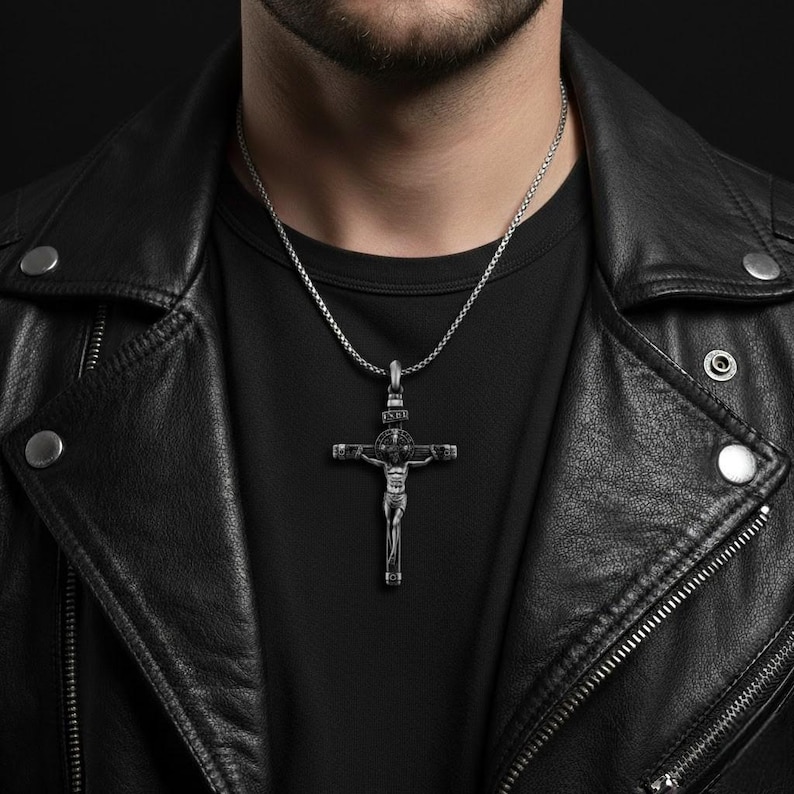 Puede incluir: Un colgante de crucifijo plateado cuelga de una fina cadena. El colgante detallado representa una figura en la cruz. El collar se lleva sobre una camiseta negra y una chaqueta de cuero negra.