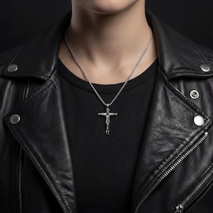 May include: A silver crucifix pendant hangs from a chain necklace. The pendant depicts a figure on a cross. The necklace is worn over a black t-shirt and a black leather jacket.