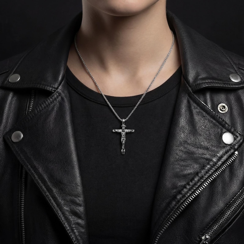 May include: A silver crucifix pendant hangs from a chain necklace. The pendant depicts a figure on a cross. The necklace is worn over a black t-shirt and a black leather jacket.