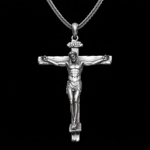 May include: A silver-colored pendant necklace featuring a detailed depiction of Jesus on the cross. The cross has the letters "INRI" above the figure. The necklace hangs on a silver chain against a black background.
