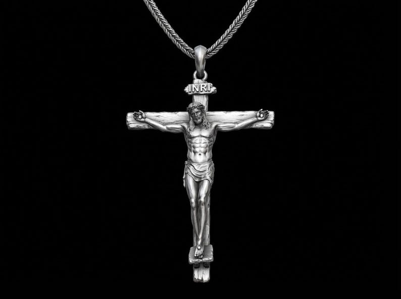 May include: A silver-colored pendant necklace featuring a detailed depiction of Jesus on the cross. The cross has the letters "INRI" above the figure. The necklace hangs on a silver chain against a black background.
