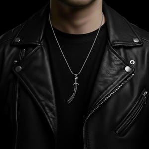 May include: Silver-toned pendant necklace featuring a curved sword design. The pendant has intricate detailing and hangs from a delicate chain. The necklace is displayed against a black background, worn over a black shirt and a black leather jacket.
