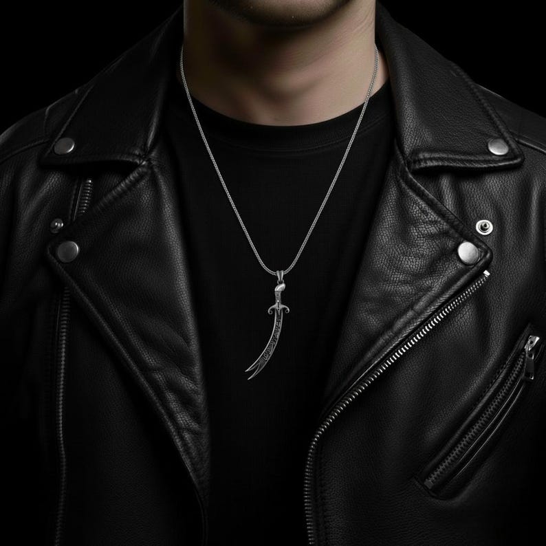 May include: Silver-toned pendant necklace featuring a curved sword design. The pendant has intricate detailing and hangs from a delicate chain. The necklace is displayed against a black background, worn over a black shirt and a black leather jacket.