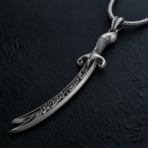May include: A silver-colored pendant necklace featuring a curved sword design. The sword has intricate detailing on the handle and blade, with engraved Arabic script. The necklace chain is a woven style, and the pendant is displayed against a dark background.