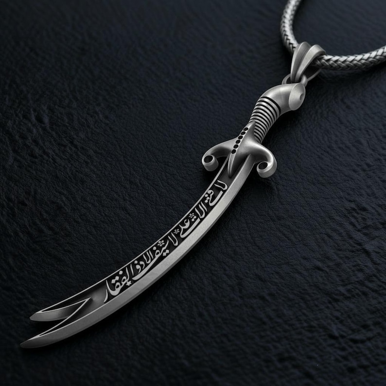 May include: A silver-colored pendant necklace featuring a curved sword design. The sword has intricate detailing on the handle and blade, with engraved Arabic script. The necklace chain is a woven style, and the pendant is displayed against a dark background.