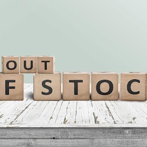 May include: A series of light brown wooden blocks arranged in two rows, with the top row spelling OUT and the bottom row spelling OF STOCK in bold black capital letters. The blocks are positioned on a white, weathered wooden surface.