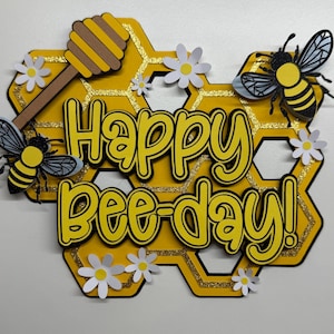 May include: A decorative wall sign featuring a yellow honeycomb background with gold glitter accents. The text reads Happy Bee-day! in black and yellow lettering. It is decorated with two black and yellow bees, a brown honey dipper, and white daisies.