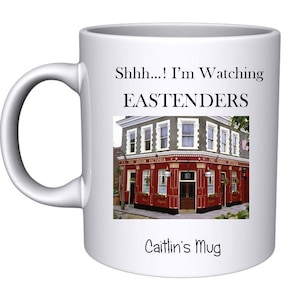 May include: White ceramic mug featuring a black and white image of the Queen Victoria pub from the TV show EastEnders. The mug has the text "Shhh... I'm Watching Eastenders" and "Caitlin's Mug" printed on it.