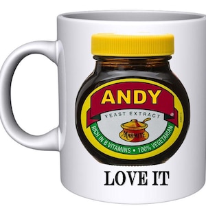 May include: White ceramic mug featuring a black and yellow jar of Marmite yeast extract. The jar has the text "ANDY YEAST EXTRACT" and "RICH IN B VITAMINS - 100% VEGETARIAN" on the label. The mug also has the text "LOVE IT" printed on the bottom.