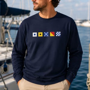 May include: A navy blue long-sleeve shirt featuring a horizontal row of five colorful nautical signal flags printed across the chest. The flags include a white square with a blue border, a yellow square with a black circle, a blue and white cross, a yellow and red flag, and a blue and white checkered square.