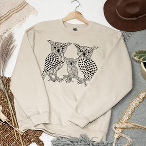May include: Cream-colored sweatshirt featuring a black graphic of three owls. The owls are stylized with dotted patterns. The sweatshirt is hanging on a wooden hanger. The background includes a brown hat, a plant, and decorative items.