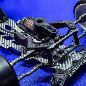 Puede incluir: Primer plano de un chasis de coche radiocontrolado negro y plateado. El chasis presenta un patrón de tejido de fibra de carbono y varios componentes mecánicos, incluido un servo con el texto "Futaba HPS GT701". Se ven las ruedas.
