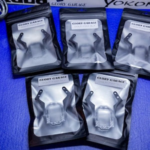 Puede incluir: Cinco bolsas de plástico negro que contienen piezas de automóviles están dispuestas sobre una superficie azul. Cada bolsa tiene una etiqueta que dice "GLORY GARAGE" y un código de producto. Las piezas parecen ser soportes para un vehículo.