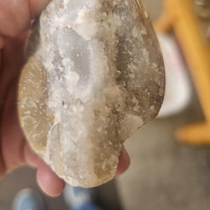 May include: A close-up of a light gray and brown agate stone with a white crystalline texture. The stone is smooth and has a rounded shape.