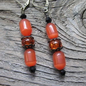 May include: Orange and brown glass bead earrings with bronze metal accents. The earrings are hanging from a wooden surface.