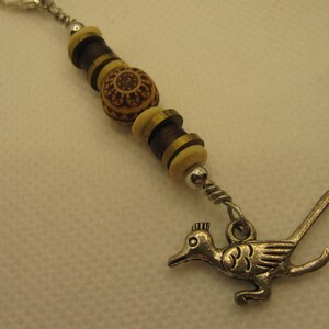 May include: A beaded keychain featuring a silver-colored roadrunner charm. The beads are a combination of dark brown, light brown, and cream-colored wood. The keychain has a silver-colored clasp.