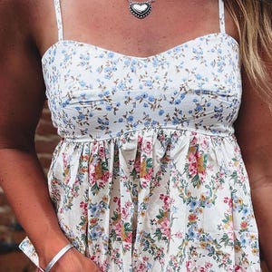 May include: A white camisole top featuring a sweetheart neckline and thin spaghetti straps. The bodice displays a small blue floral pattern, while the lower section features a larger, vibrant floral print with pink, yellow, and blue flowers.