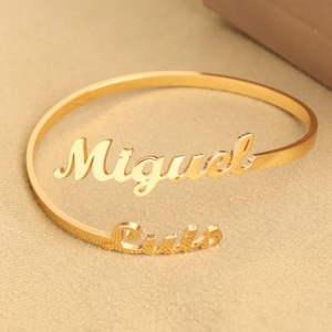 May include: A gold-coloured cuff bracelet with the names "Miguel" and "Rocio" in a cursive font. The open-ended bracelet rests on a beige surface. The bracelet is a piece of jewellery.