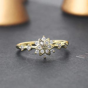 May include: A gold-colored ring featuring a snowflake design. The ring has a delicate band and is adorned with small, clear gemstones. The snowflake is the focal point, with multiple gemstones set within its intricate pattern. The ring is set against a dark background.