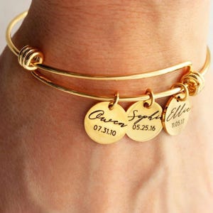 May include: Gold-coloured bangle bracelet with three round charms. Each charm is engraved with a name and date. The bracelet is worn on a wrist.