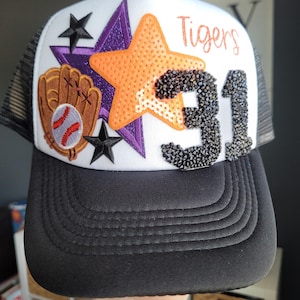 May include: White baseball cap with a black mesh back. The front of the cap features a purple and orange star, a baseball glove and ball, and black rhinestones spelling out "Tigers 31".