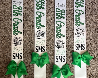 Puede incluir: Cuatro bandas de satén blanco con texto en purpurina verde que dice 8th Grade, Cheer, 26-27 y SMS, junto con los nombres Raelyn, Maycee, Amelia y Lily. Cada banda está decorada con un lazo de tela verde brillante en el borde inferior.