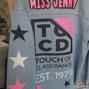 Puede incluir: Una chaqueta vaquera azul claro con el texto "Miss Jenny" y "Touch of Class Dance Est. 1979" en la espalda. La chaqueta tiene estrellas de purpurina negras, blancas y rosas en las mangas.