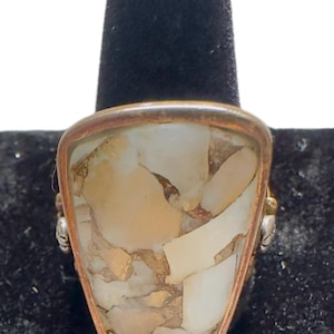 Può includere: Un anello con un grande inserto a forma di scudo composto da frammenti di pietra color crema, marrone chiaro e bianco con venature color rame metallizzato. L'inserto è incastonato in una cornice di metallo liscio color rame lucido.