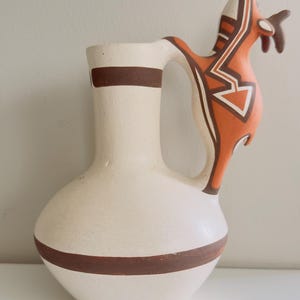 May include: A cream-colored ceramic pitcher featuring a rounded base and a tall neck with a brown horizontal band. The handle is shaped like a stylized bird, painted in orange, white, and brown geometric patterns, extending from the neck to the body.