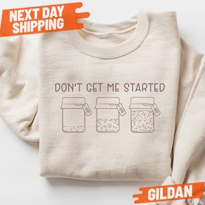 May include: Sand-colored Gildan sweatshirt with the phrase "DON'T GET ME STARTED" and three jar illustrations. Each jar has a different fill level and a small tag. The sweatshirt has an orange "NEXT DAY SHIPPING" and "SAND" tag.