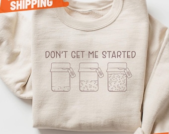 May include: Sand-colored Gildan sweatshirt with the phrase "DON'T GET ME STARTED" and three jar illustrations. Each jar has a different fill level and a small tag. The sweatshirt has an orange "NEXT DAY SHIPPING" and "SAND" tag.
