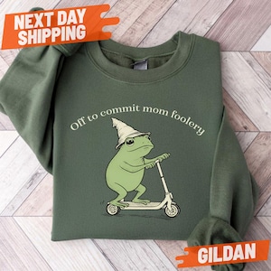 May include: Military green sweatshirt with a cartoon frog riding a scooter and wearing a wizard hat. The text "Off to commit mom foolery" is printed above the frog. The sweatshirt is a Gildan brand.