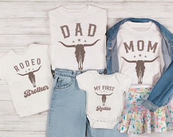 May include: A collection of white t-shirts and a onesie with rodeo-themed graphics. The shirts feature the words "DAD," "MOM," and "Brother," along with a bull skull design. A baby onesie says "My First Rodeo."