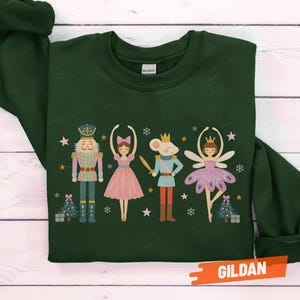 May include: Forest green sweatshirt featuring a Nutcracker-themed design. The graphic includes a nutcracker, a ballerina, a mouse king, and a fairy, with Christmas trees and stars. The brand name "Gildan" is visible.