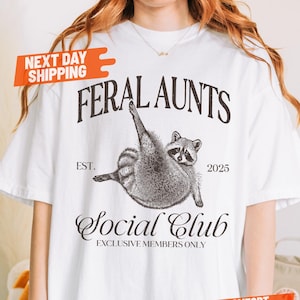 May include: White t-shirt featuring a raccoon illustration with the text "FERAL AUNTS Social Club". The design includes the year "2025" and the words "EXCLUSIVE MEMBERS ONLY". The shirt is a comfort color.