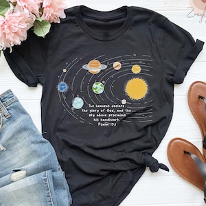 May include: Black t-shirt featuring a solar system design with planets and the sun in yellow, orange, blue, and green. The text "The heavens declare the glory of God, and the sky above proclaims his handiwork. Psalm 19:1" is printed below the design.