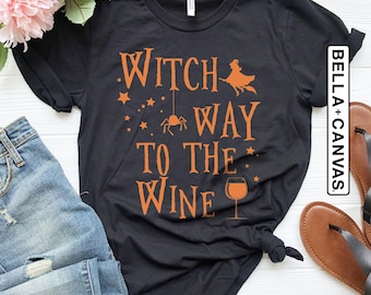 May include: Black t-shirt with orange text that reads "Witch Way To The Wine" with a witch on a broom, stars, a spider, and a wine glass. The shirt is styled with blue jeans, sandals, and pink flowers.