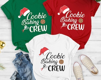 May include: Three short-sleeved t-shirts in green, red, and white, each with the text "Cookie Baking Crew" and a Santa hat graphic. A pair of blue jeans and sneakers are also in the image.