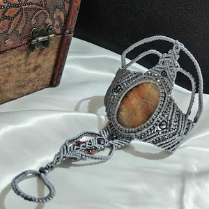 Può includere: Un braccialetto in macramè grigio con una grande pietra ovale di colore arancione e marrone. Il braccialetto ha un anello per il dito ed è decorato con piccole perline nere.