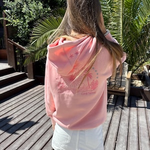 Könnte beinhalten: Ein rosa Hoodie mit einem Aufdruck eines Palmenbaums, einer Sonne und eines Korallenriffs auf der Rückseite. Der Text "Coral Sunset" ist unter dem Aufdruck gedruckt.