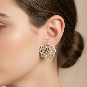 May include: Gold-toned stud earrings with a floral design. The earrings feature a central clear gemstone surrounded by red and clear accents, and a border of small white beads. A classic jewelry piece.