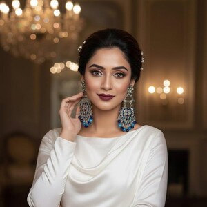 May include: A woman wearing large, ornate silver earrings with blue and silver bead accents. She is wearing a white satin dress with a cowl neckline. The background is softly lit with a chandelier and warm tones.