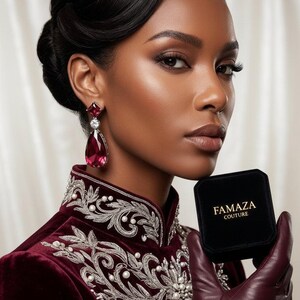 May include: A deep burgundy velvet high-neck garment featuring intricate silver embroidery and white pearl accents. The piece is paired with a large teardrop-shaped red gemstone earring and a black velvet jewellery box labelled FAMAZA COUTURE.