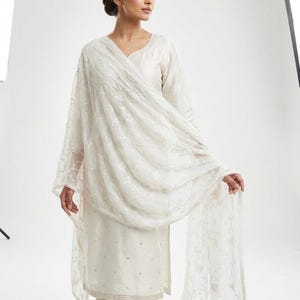 May include: A cream-coloured salwar kameez ensemble with embroidery. The outfit includes a long-sleeved tunic, matching trousers, and a sheer dupatta. The dupatta is draped over the shoulders and features intricate floral embroidery. The outfit is worn by a person.