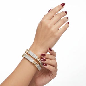 May include: Two gold-toned bangles featuring a central row of rectangular clear stones flanked by two rows of small pavé-set clear stones. The bracelets have a metallic finish and a structured design, showcasing a repeating pattern of baguette-cut gems.